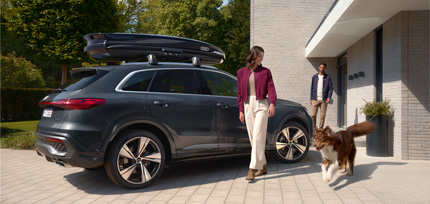 Audi SUV mit Dachbox steht vor einem Haus, w&auml;hrend zwei Personen mit einem Hund vorbeigehen.